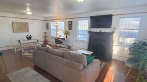 Ein Wohnzimmer mit beiger Couch, einem Esstisch mit Stühlen, einem Kamin und großen Fenstern mit Jalousien.