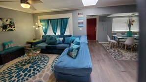 Ein Wohnzimmer mit einer Couch, einem Couchtisch und einem gemusterten Teppich. Es gibt eine Essecke mit einem Tisch und Stühlen.