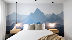 Une chambre à coucher comprenant un lit, deux tables de chevet, une commode, une fresque murale représentant des montagnes et des lampes suspendues.