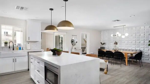 Une cuisine moderne avec des meubles blancs, un îlot en marbre, des luminaires suspendus et un coin repas comprenant une table et des chaises en bois.