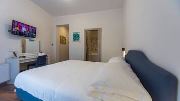 A hotel room with a bed, a desk with a computer, a TV mounted on the wall, and a door leading to another room.