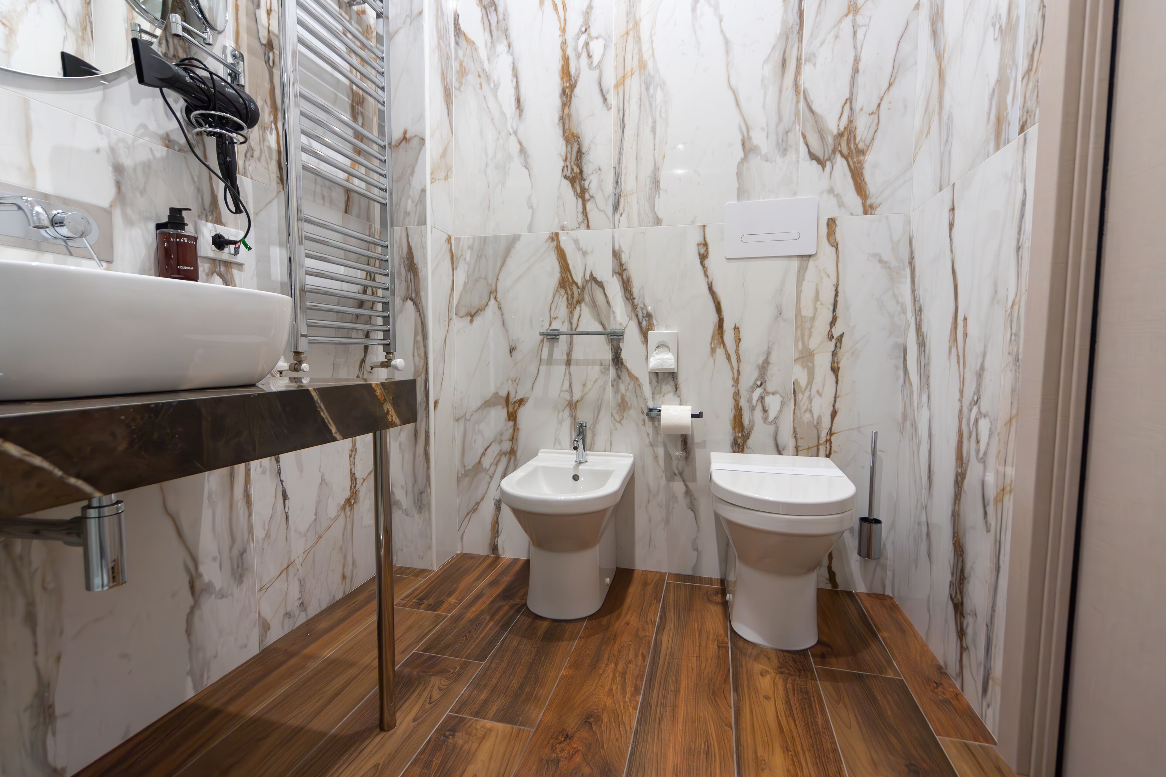 A modern bathroom with a marble backsplash, a wooden floor, a sink, a bidet, and a toilet.