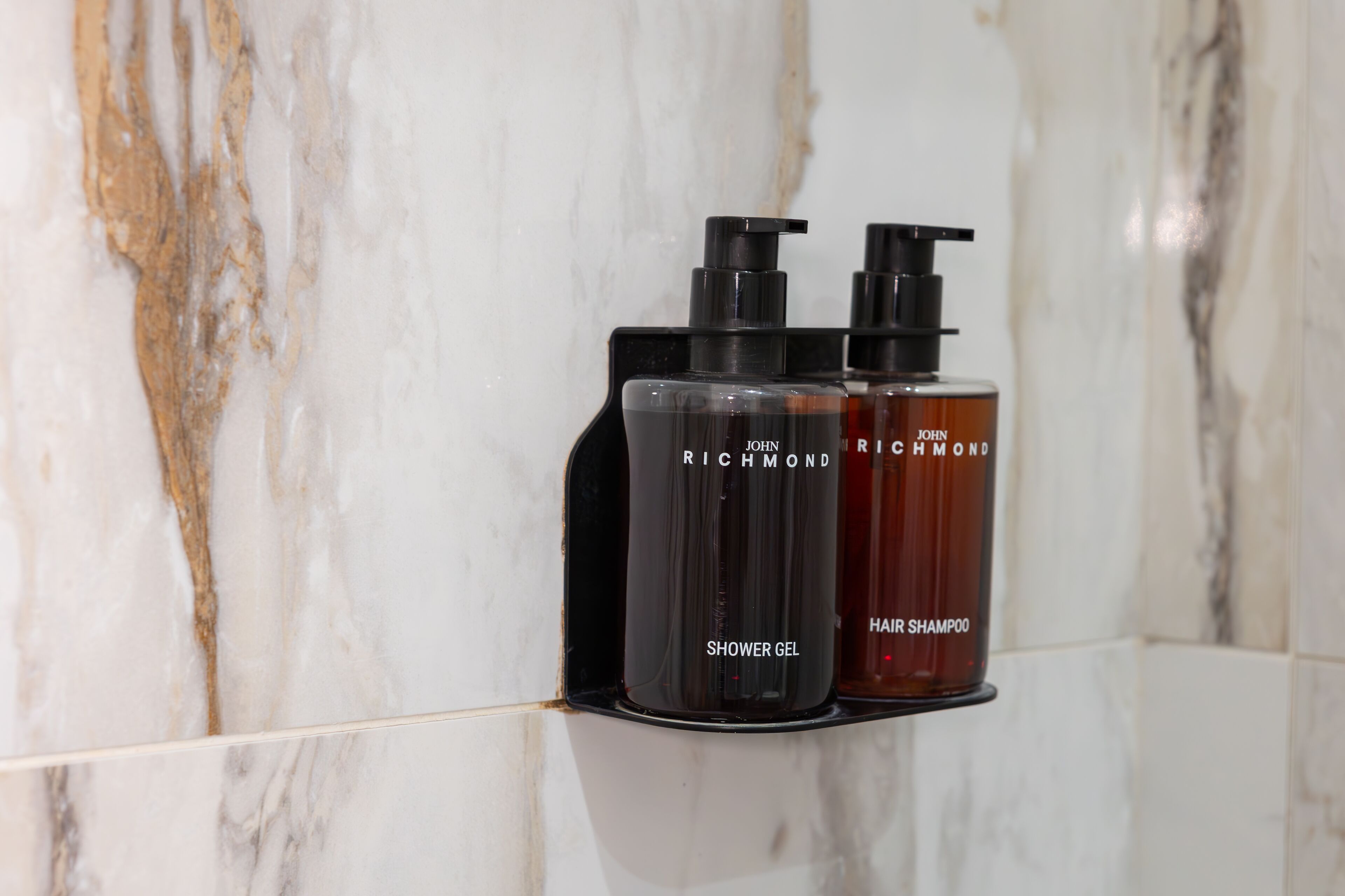 Deux flacons de gel douche et de shampoing John Richmond, posés sur un porte-savon noir devant un mur en marbre.
