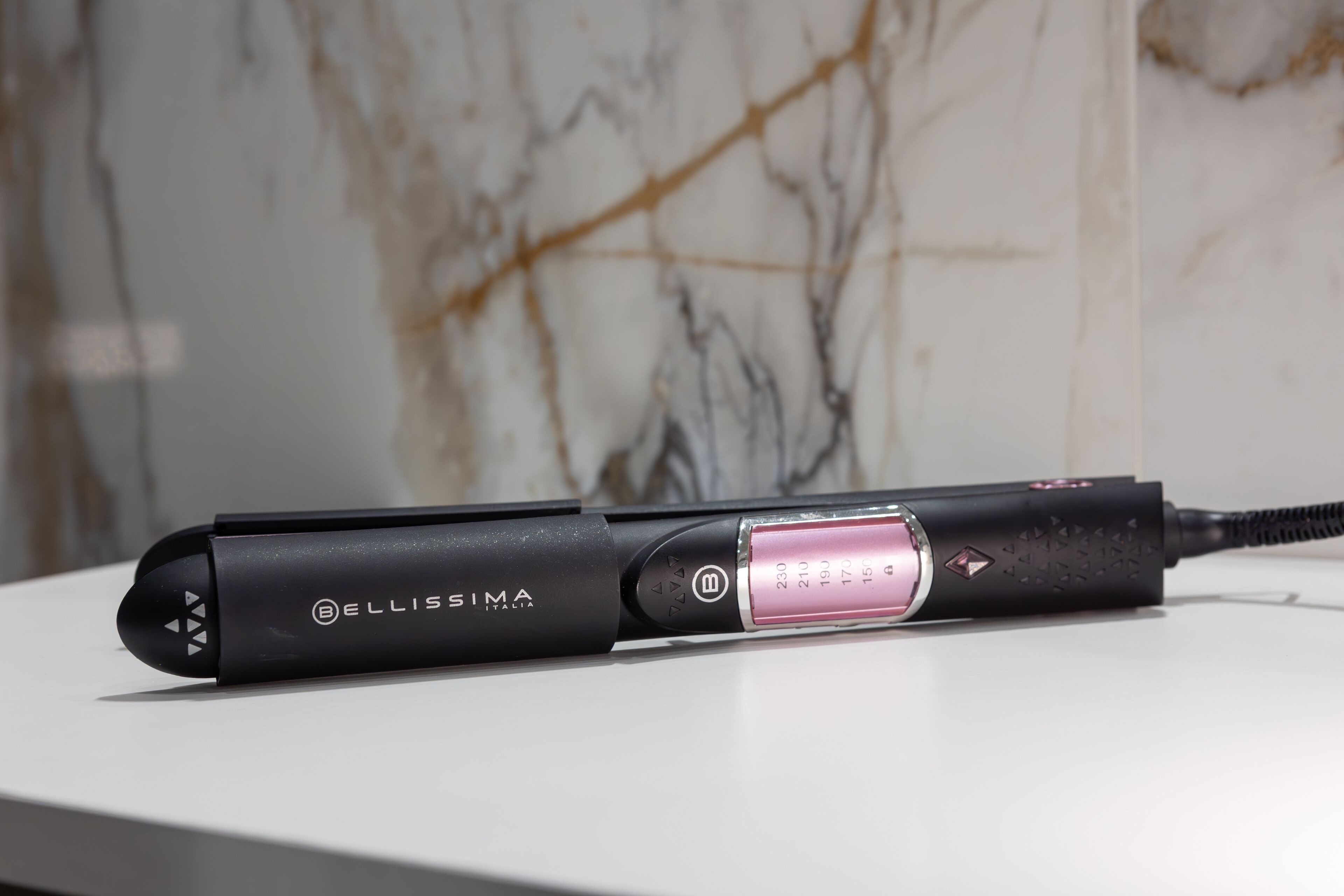 Una plancha para el cabello negra, con un detalle rosa, sobre una superficie blanca.