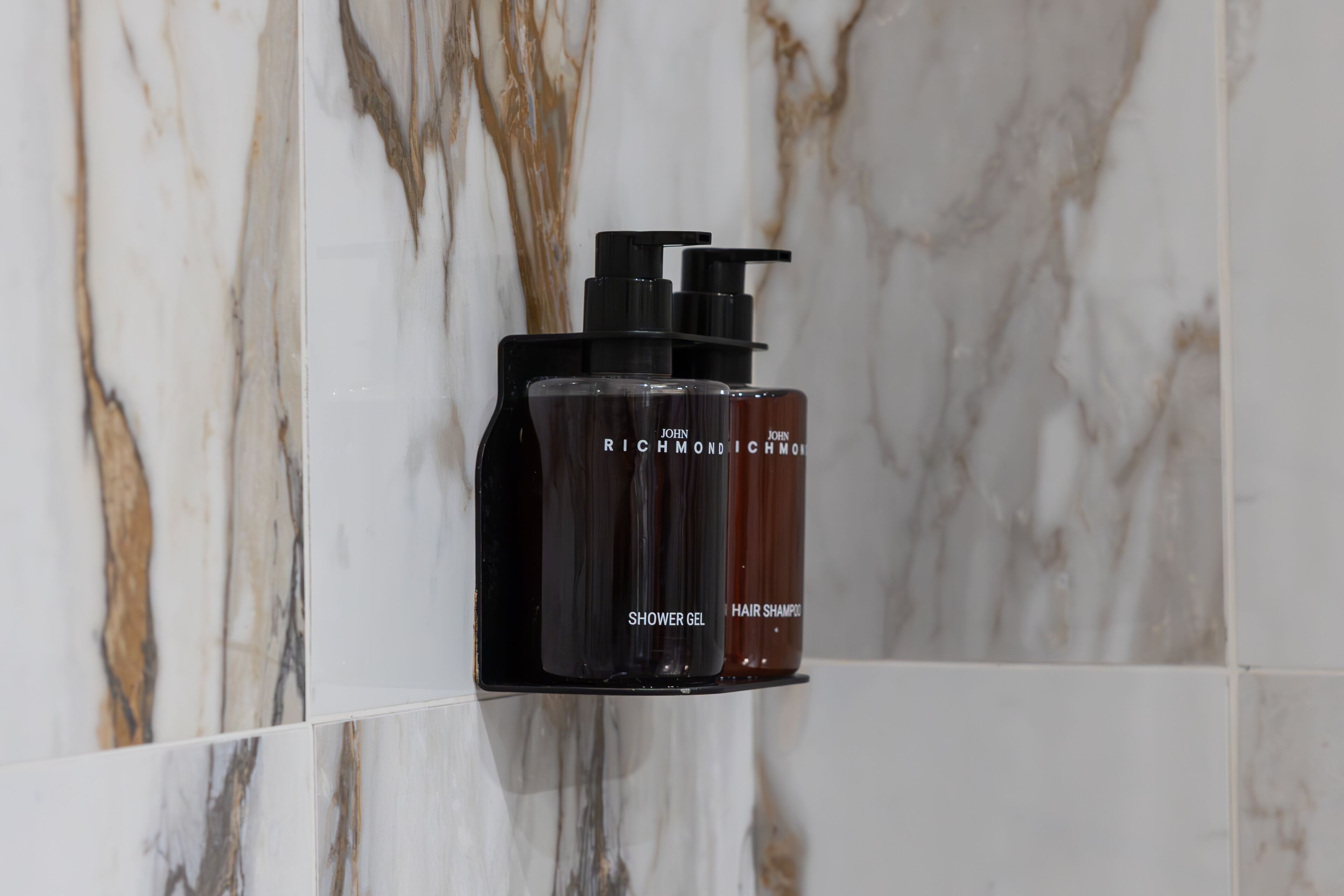 A bathroom with marble walls and a shelf holding a black bottle labeled "Shower Gel" and a brown bottle labeled "Hair Shampoo."