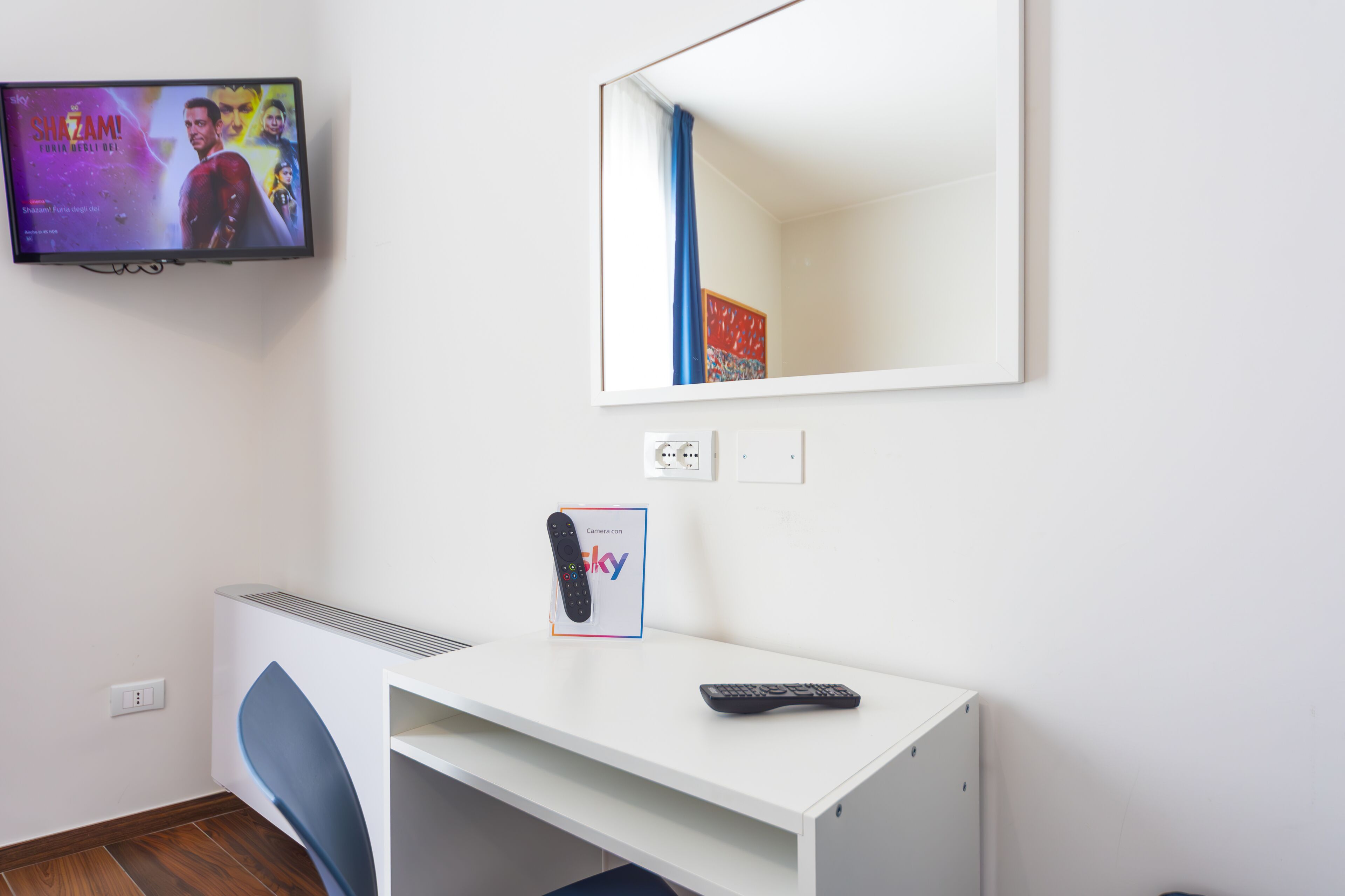 Une chambre d’hôtel équipée d’un téléviseur à écran plat diffusant un film, d’un bureau avec une télécommande, d’un miroir et d’une petite étagère sur laquelle se trouve un appareil photo.