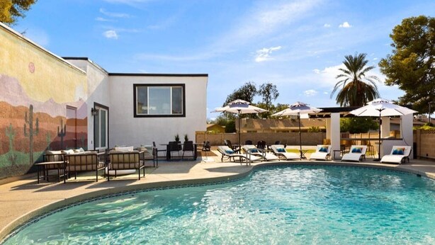 A modern pool area with lounge chairs and umbrellas, a mural on the wall, and a clear blue sky. - Heated Pool, Lavish Yard! - The Desert Wanderer (Scottsdale)