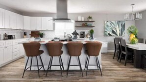 A modern kitchen with white cabinets, a central island with bar stools, and a dining area with a large painting. - Heated Pool, Lavish Yard! - The Desert Wanderer (Scottsdale)