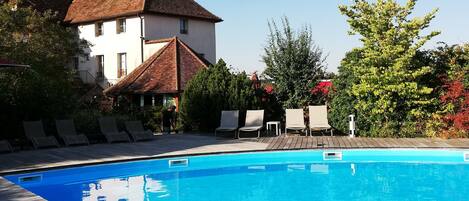 Piscina com deck de madeira, cadeiras de descanso e um edifício com telhado vermelho ao fundo.