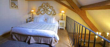 A bedroom with a large bed, wooden beams, two bedside tables with lamps, a small decorative piece on the wall, and a staircase with lights.