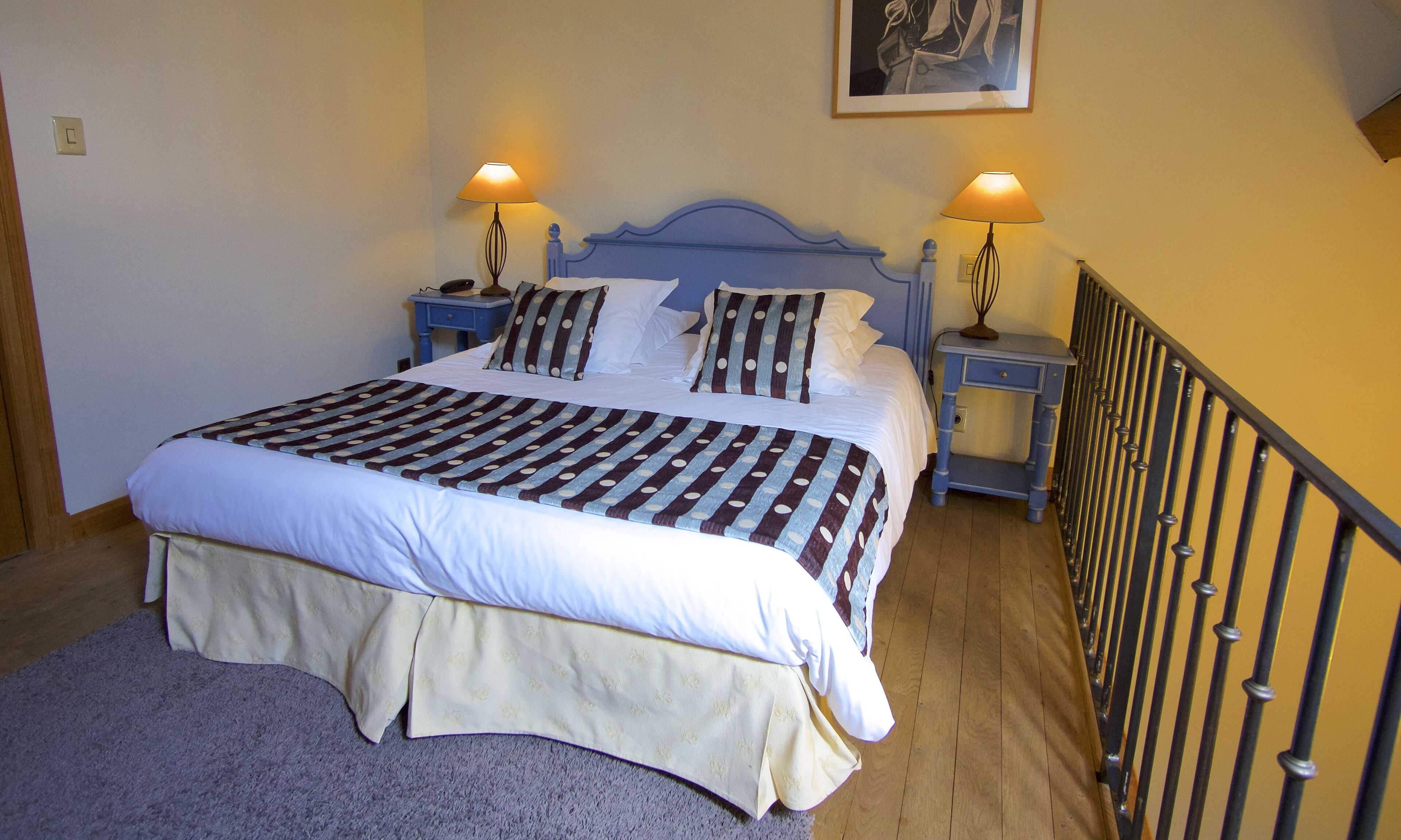 Cama bem arrumada com colcha listrada, duas luminárias de cabeceira e mesa de apoio com telefone.