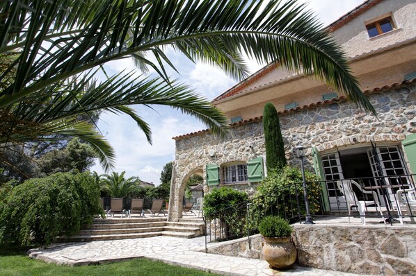 A stone building with a staircase leading to a covered entrance, surrounded by greenery and outdoor seating.
