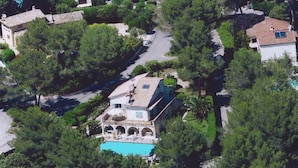 Eine Vogelperspektive zeigt eine Wohngegend mit einem markanten Haus, das einen Swimmingpool hat, umgeben von üppigem Grün und weiteren Häusern.