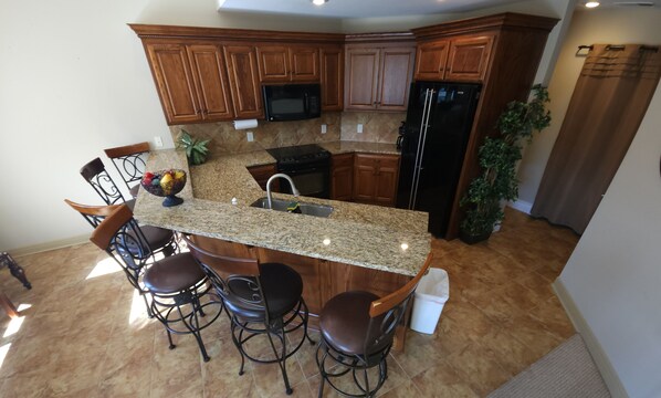 A kitchen with granite countertops, wooden cabinets, a stainless steel refrigerator, a microwave, and a sink. There are four bar stools and a potted plant. - 0ver 7,000 Sq ft of tranquil relaxation  (Shawnee)