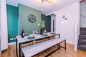 Un coin repas avec une table en bois dressée pour quatre personnes, un mur vert, une horloge et un escalier.