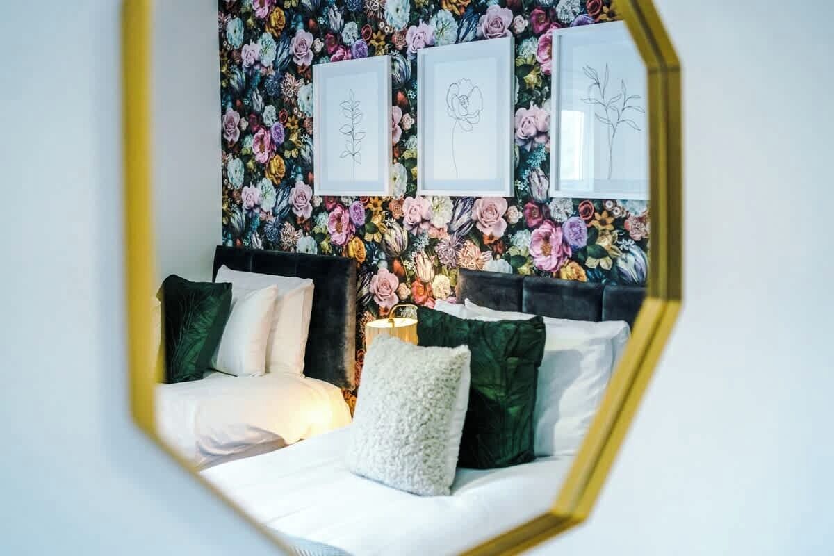 A bedroom with a floral wallpaper, a bed with white bedding and green pillows, and framed botanical drawings on the wall.