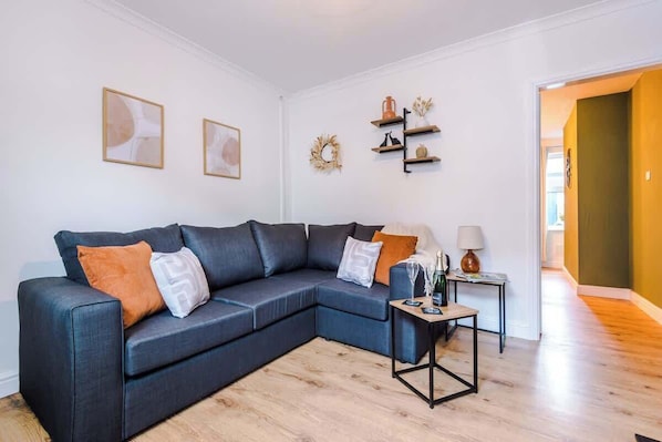 Un salon moderne avec un canapé d’angle bleu foncé, une petite table d’appoint et une étagère murale agrémentée d’objets décoratifs.