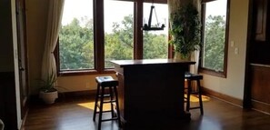 A room with a wooden floor, a bar area with stools, a hanging light fixture, and a window with a view of trees. - Peaceful Oasis (Shawnee)