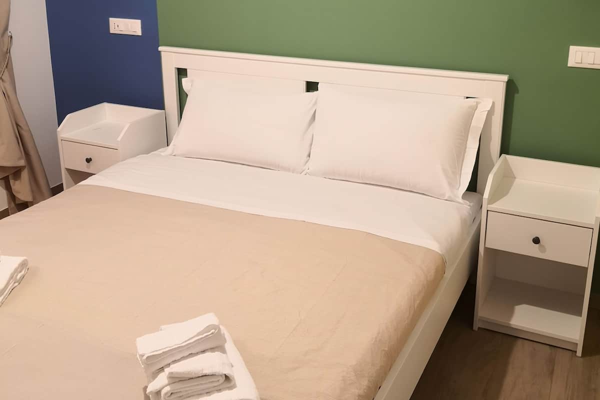Ein ordentlich bezogenes Bett mit weißen Leinentüchern und einem gefalteten Handtuch am Fußteil. Zwei Nachttische, jeweils mit Lampe und Schublade. Die Wand hat einen blauen und grünen Akzent.