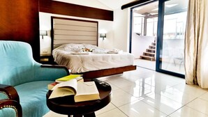 A hotel room with a bed, a blue armchair, a small table with books, and a view of the outside through a large window. - Galini Private Pool Suites with Sea View (Chania)