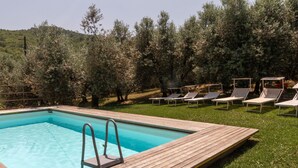 Ein rechteckiger Pool mit Holzdeck und Metallleiter, umgeben von einer Rasenfläche mit mehreren Liegestühlen und einer Baumreihe im Hintergrund.