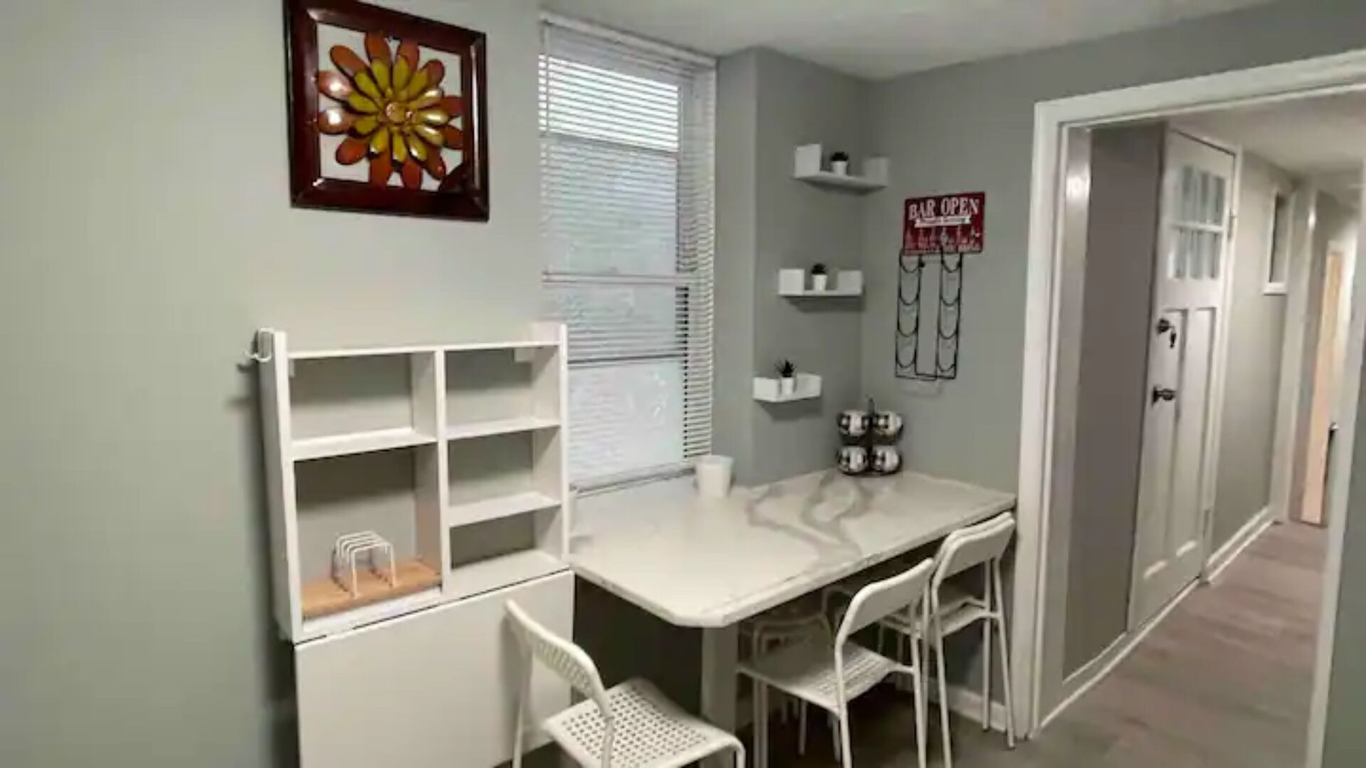 A room with a white table, two chairs, a window with blinds, a door, and a 'BAR OPEN' sign.