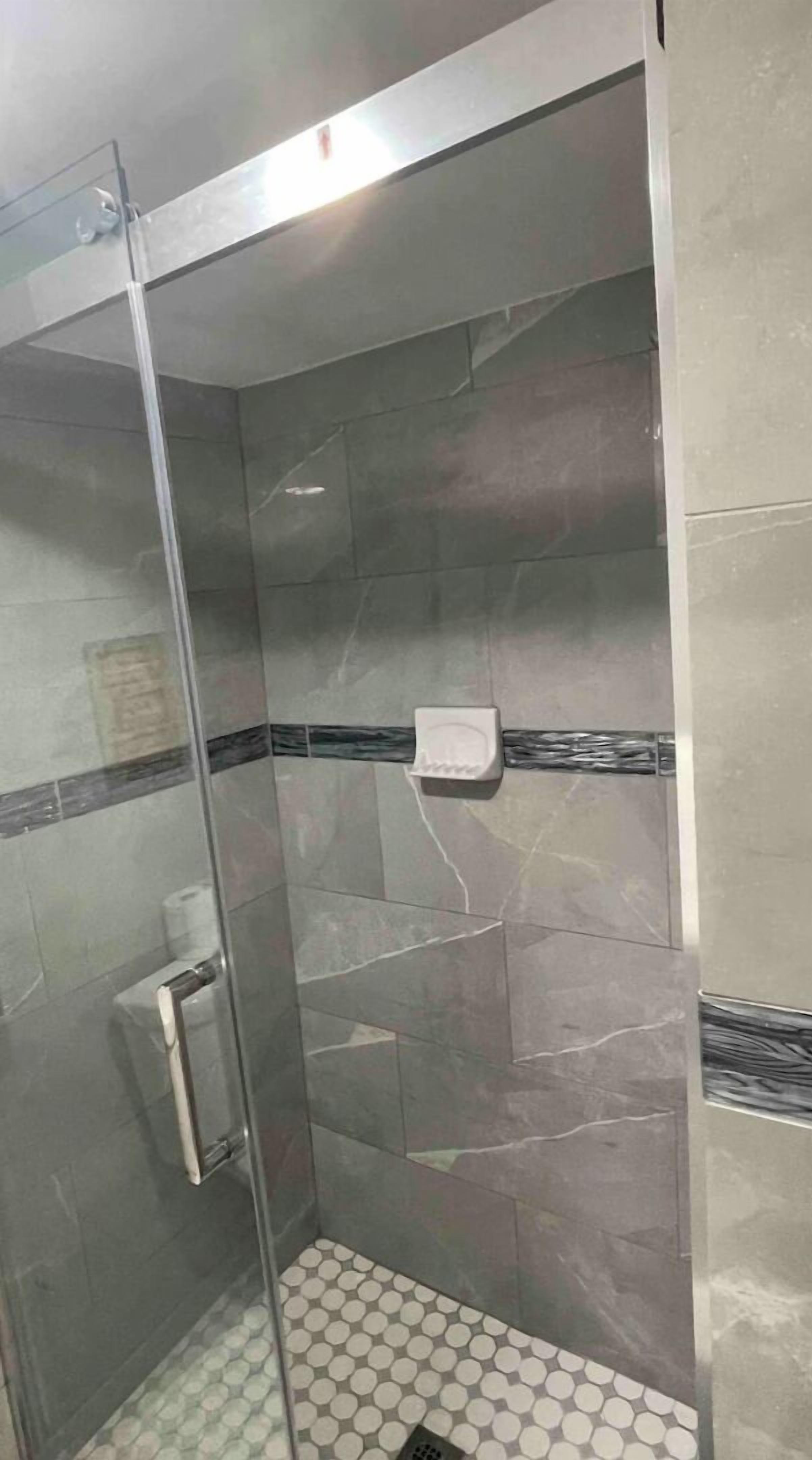 Un área de ducha con puerta de vidrio, paredes con azulejos y piso estampado.