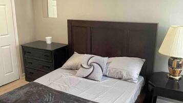 Un letto ben rifatto con una testata scura, un comodino con una lampada e un piccolo comò con una tazza sopra.