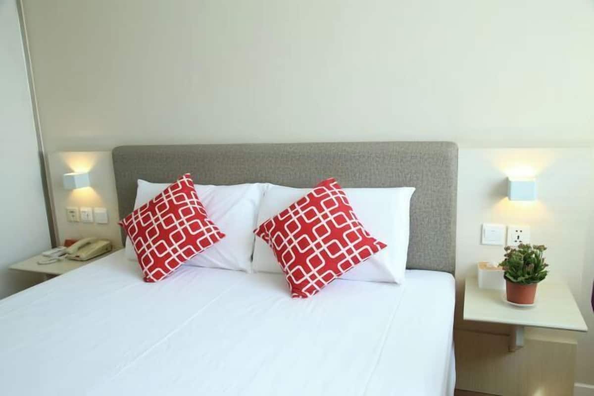 Une chambre d’hôtel avec un lit, deux oreillers à motifs rouges et blancs, une table de chevet avec un téléphone et une plante, et des lampes fixées au mur.