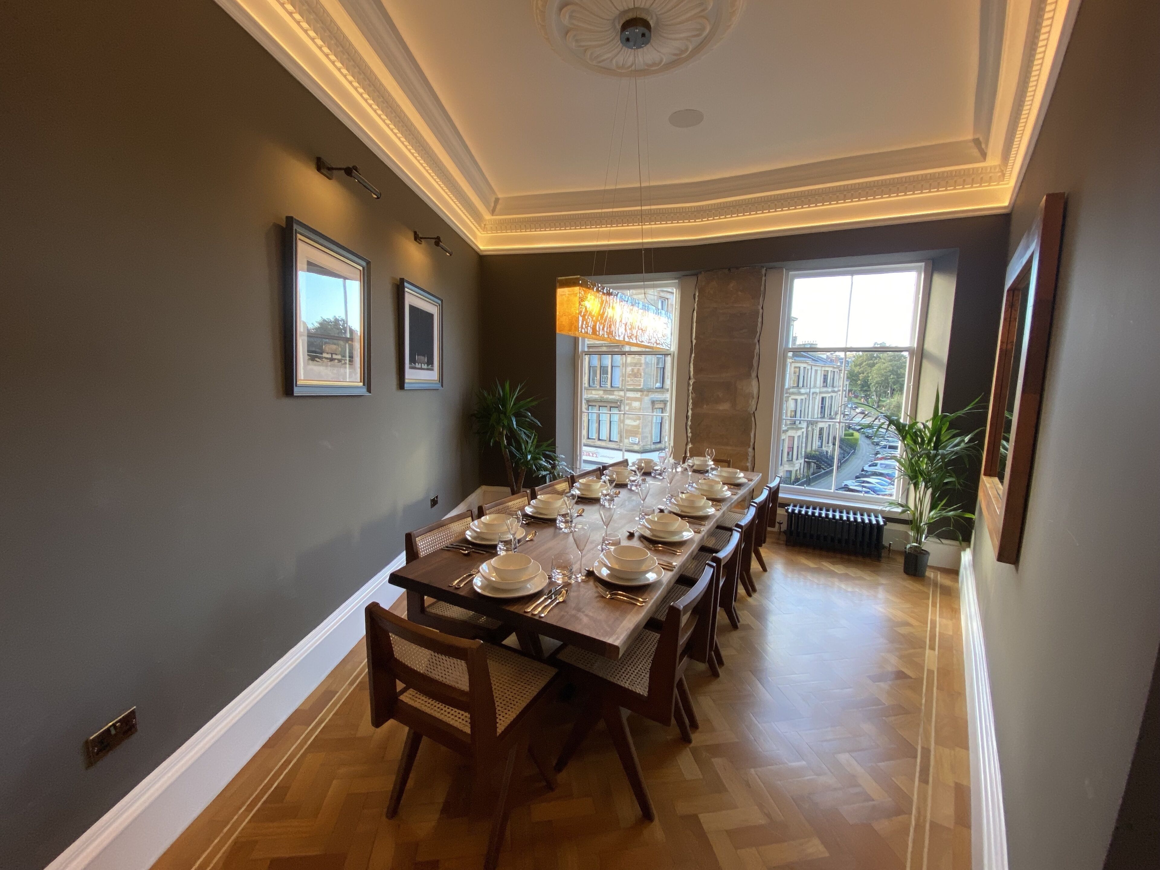 A dining room with a long table set for six, framed pictures on the wall, and a view of buildings through large windows.