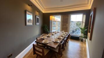 A dining room with a long table set for six, framed pictures on the wall, and a view of buildings through large windows.