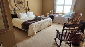 Ein Schlafzimmer mit einem großen Bett, einem kleineren Bett, einem Schaukelstuhl, einer hölzernen Raddekoration und einem Fenster mit Vorhängen.