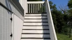 Eine Treppe mit weißen Stufen führt zum Eingang einer Hausfassade. Die Tür ist blau, das Geländer weiß.
