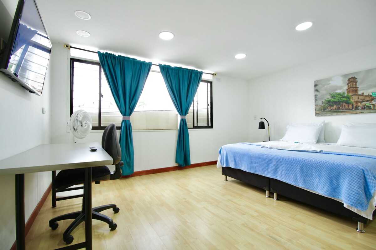 Une chambre d’hôtel équipée d’un lit, d’un bureau, d’une chaise, d’un ventilateur et d’une fenêtre avec des rideaux.