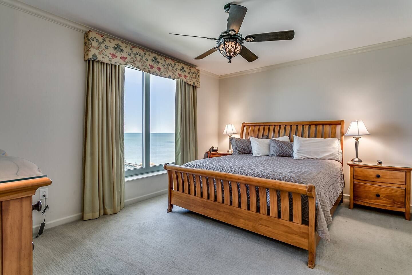 A bedroom with a wooden bed, two bedside tables with lamps, a ceiling fan, a window with curtains, and a view of the ocean.