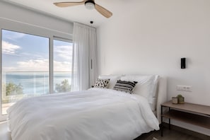A bedroom with a large bed, a ceiling fan, a nightstand, and a view of the sea.