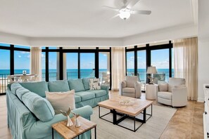 A spacious living room with a large window offering a view of the ocean, a ceiling fan, and a seating area with a sofa and armchairs.