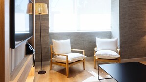 A modern hotel room with a large window, two wooden chairs, a black table, a flat-screen TV mounted on the wall, and a floor lamp. - THE Apartment by Assethoteles (Torrejón de Ardoz)