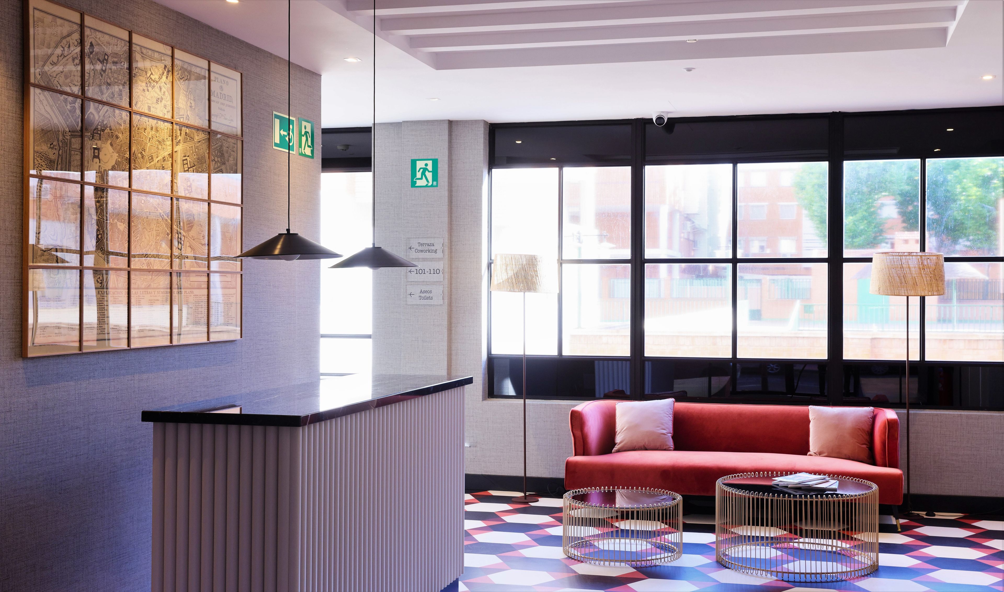 A hotel lobby with a reception desk, a red sofa, two round coffee tables, a large window with a view, and directional signs.