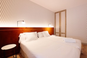 A hotel room with a large bed, two bedside lamps, a wooden headboard, a small table, and a wardrobe. - THE Apartment by Assethoteles (Torrejón de Ardoz)