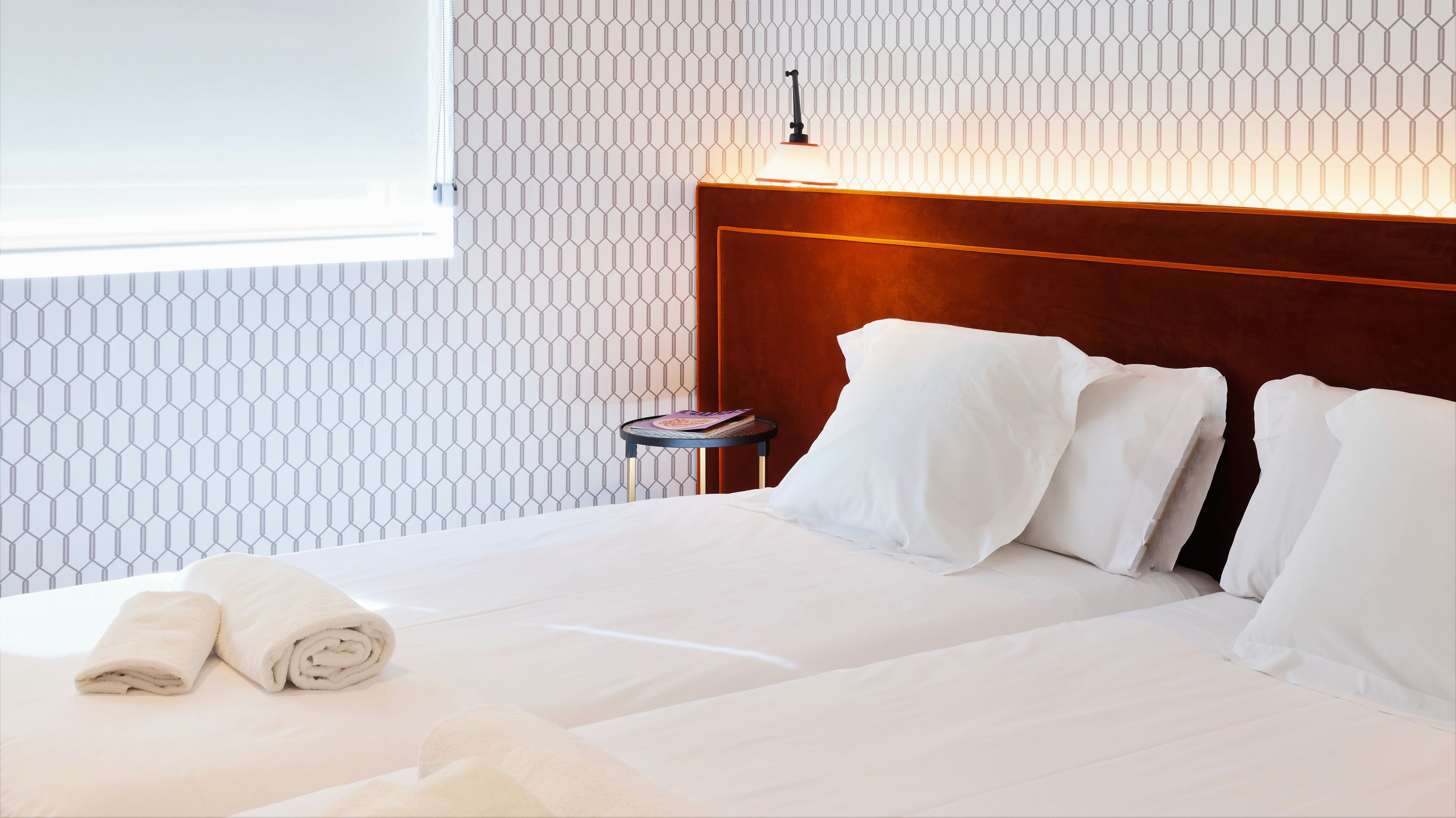 A neatly made bed with white linens and pillows, a bedside table with a book, and a wall with a geometric pattern.