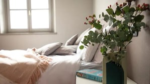 A neatly made bed with a white blanket and pillows, a bedside table with a book and a potted plant, and a window providing natural light. - Classic Double with panoramic view (Toscana)