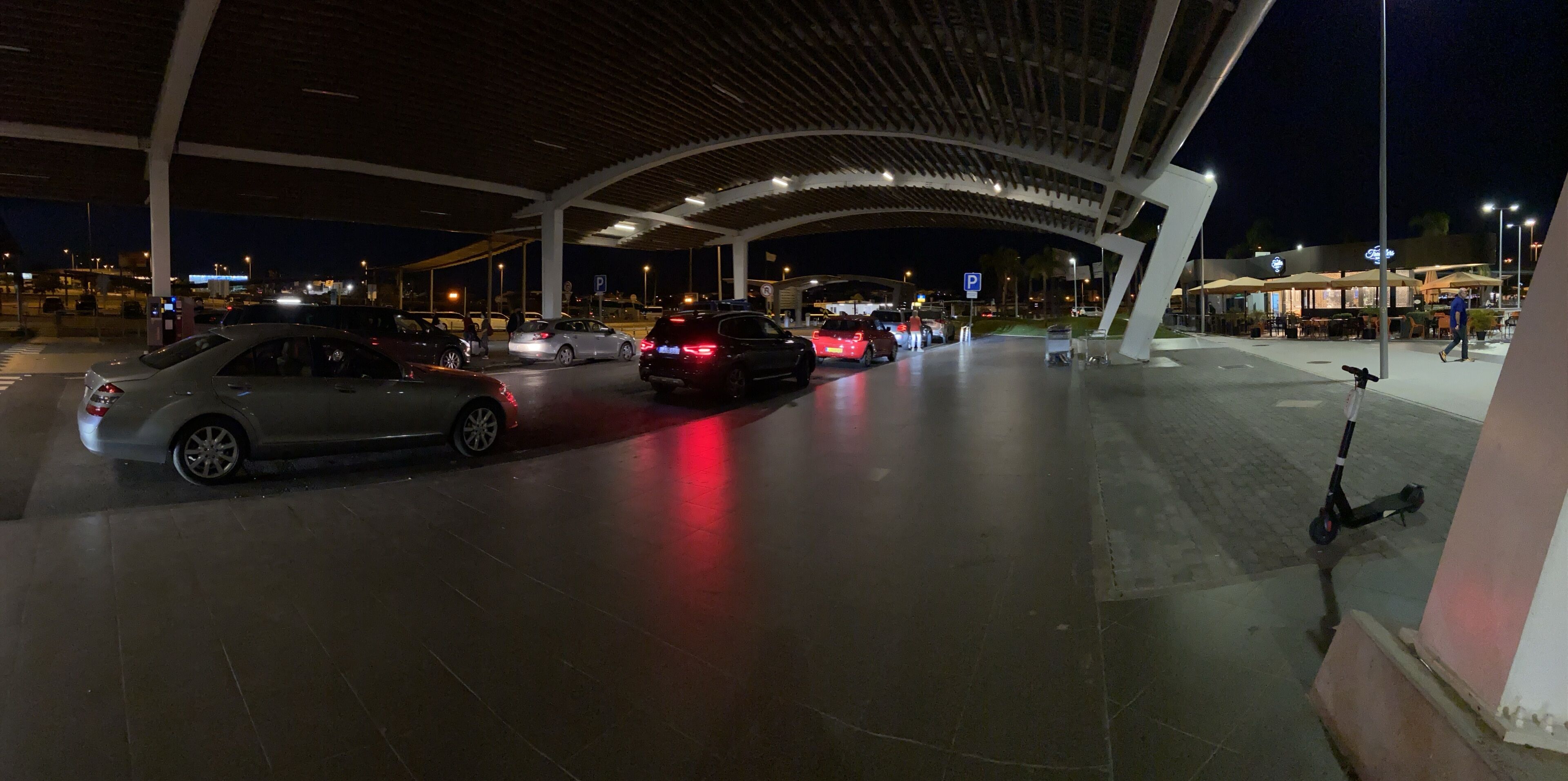 Un área de estacionamiento con coches y una moto, una estructura cubierta y un edificio con terraza al fondo.