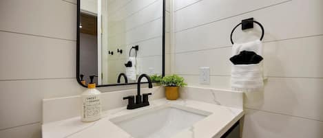Un baño con un lavamanos blanco, grifo negro y un espejo. Hay un perchero con una toalla blanca y una toalla negra. Una pequeña planta en maceta está sobre el mostrador.