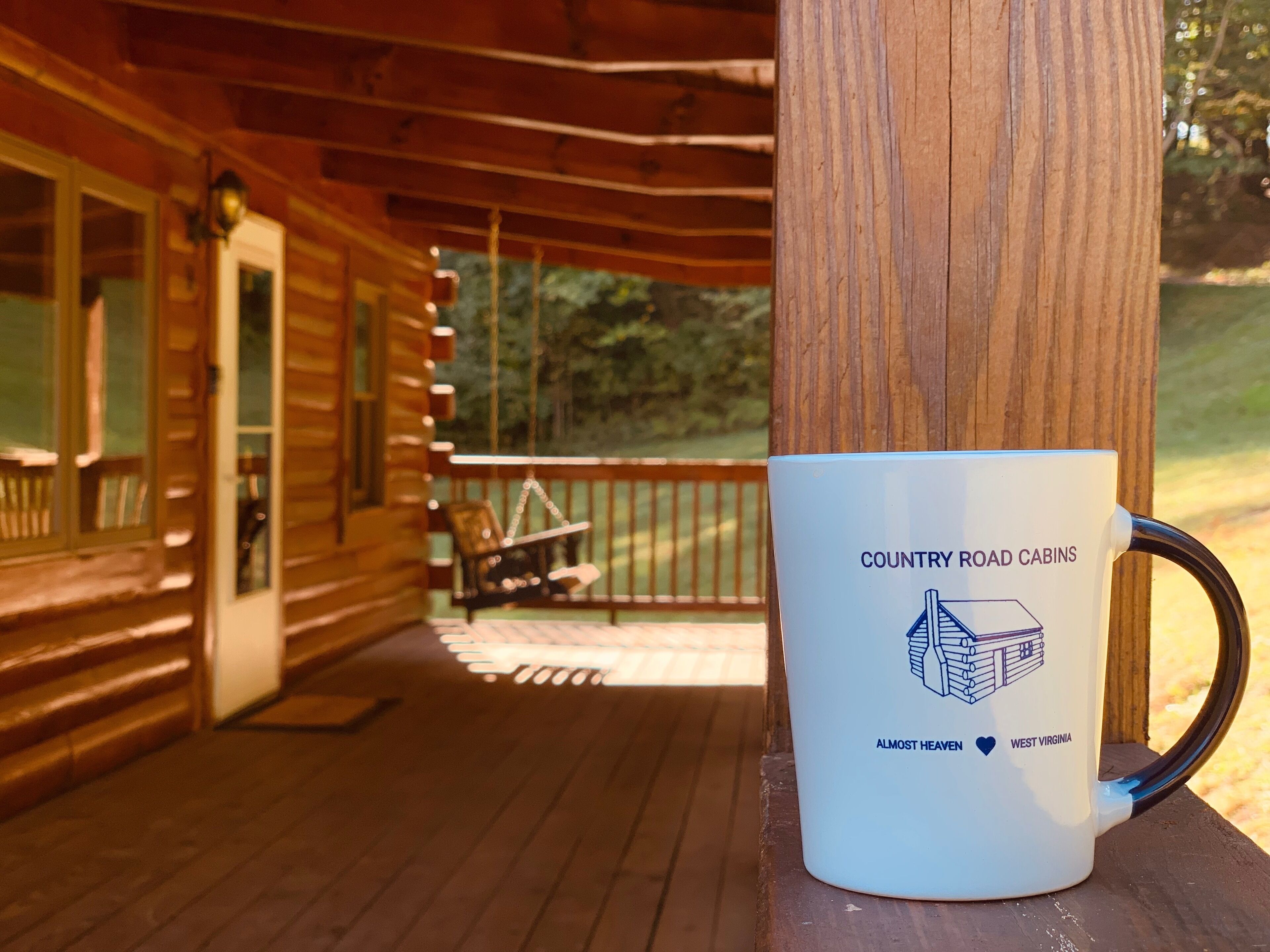 Una taza blanca con el texto 'Country Road Cabins' y una ilustración de una cabaña, sobre una superficie de madera.