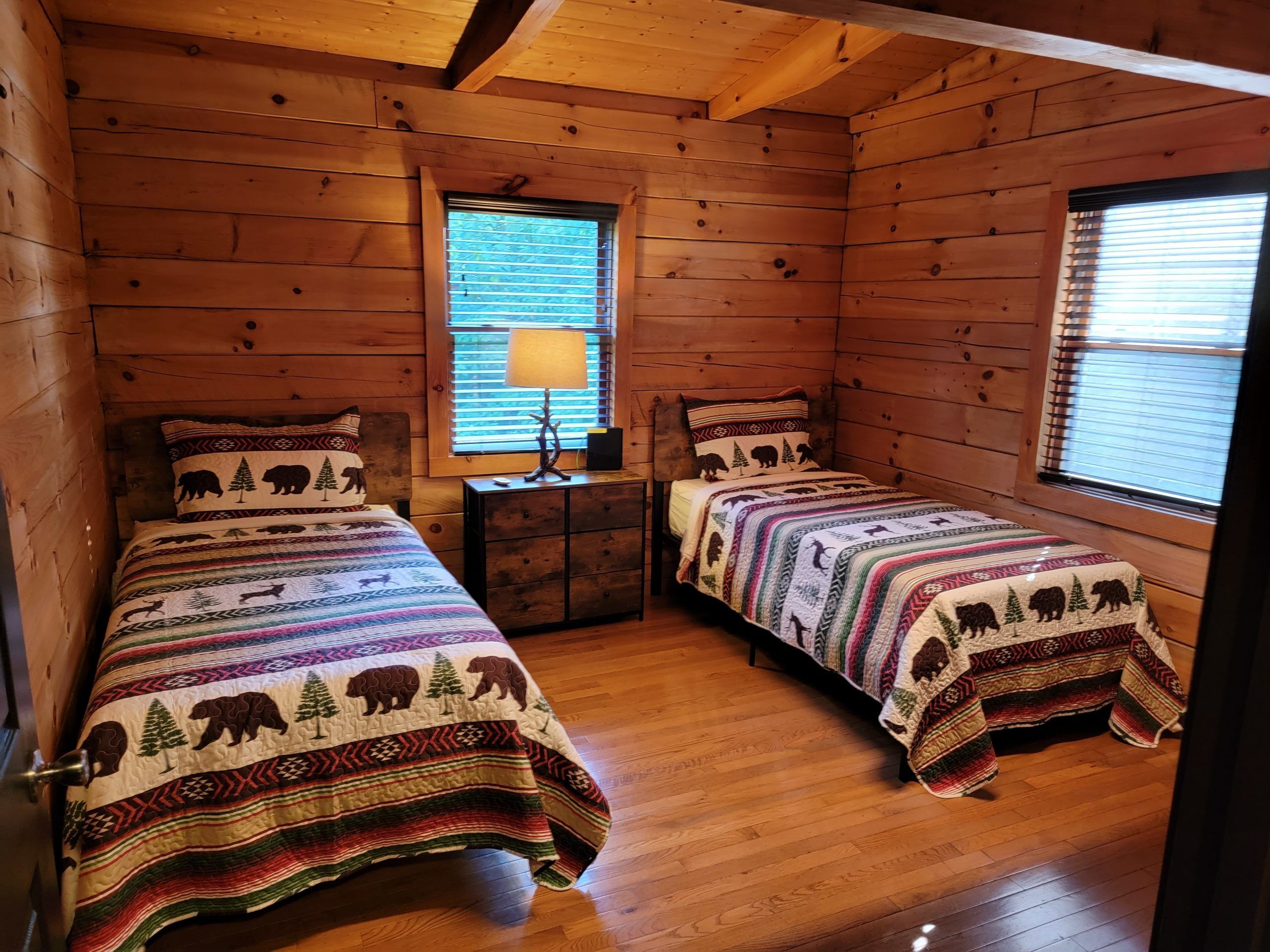 Ein Blockhaus-Zimmer mit zwei Betten, Holzwänden, einem Fenster mit Jalousien und einem Nachttisch mit Lampe.
