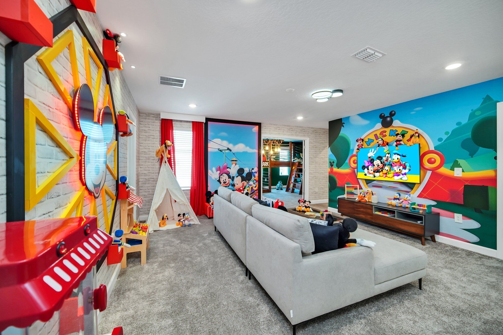 Ein modernes Wohnzimmer mit einem grauen Sofa, einem Spielbereich mit einer Rutsche und einem großen Fernsehbildschirm, der Zeichentrickfiguren zeigt.