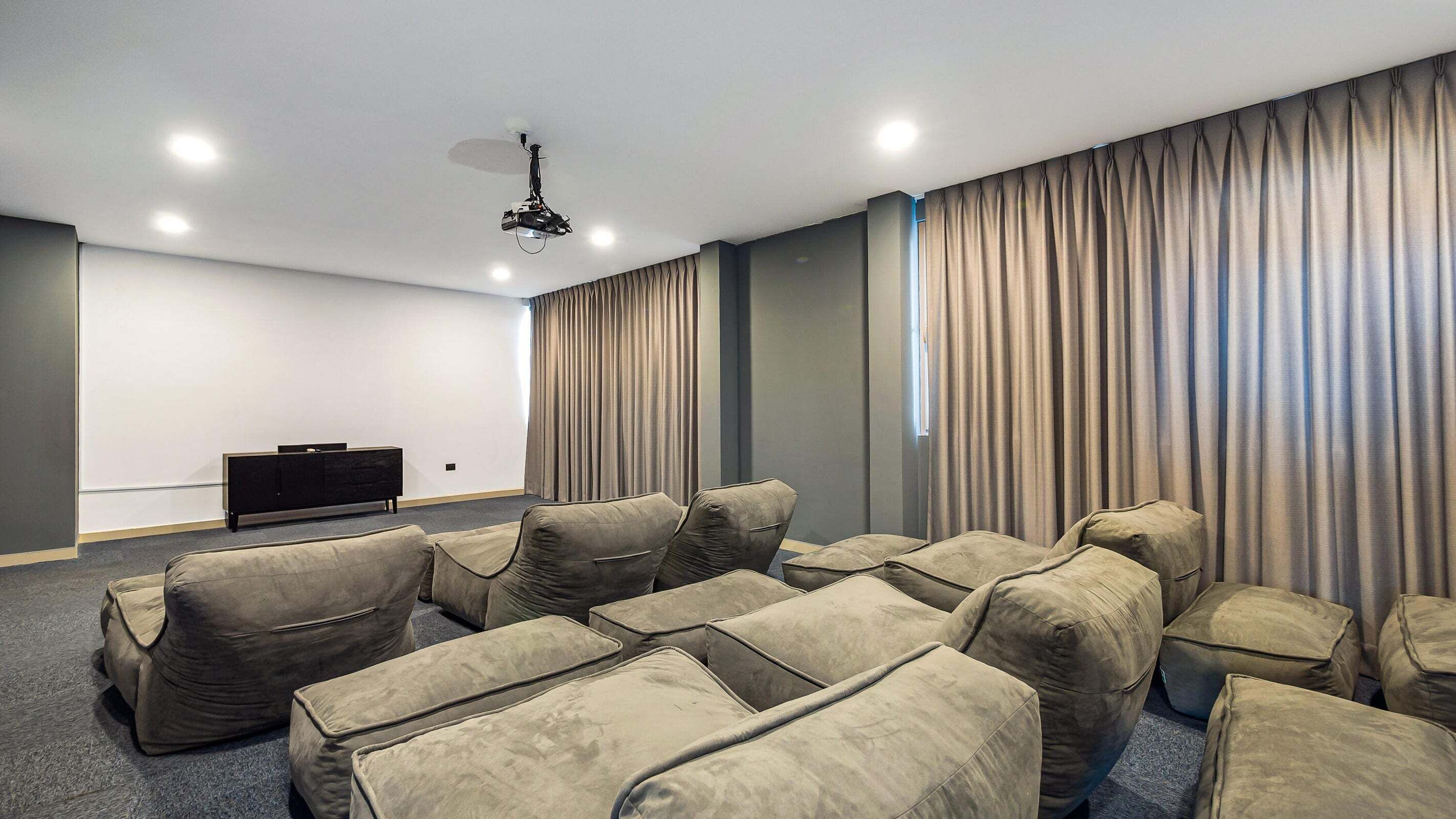 Un cine con hileras de asientos color beige acolchados, una pantalla oscura y cortinas en las ventanas.