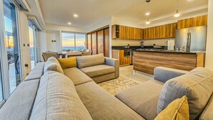 Een moderne woonkamer met een ruime hoekbank, een keuken met houten kasten en uitzicht op de ondergaande zon door grote ramen.