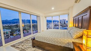Een slaapkamer met een groot bed, een nachtkastje en een stadsgezicht door grote ramen.